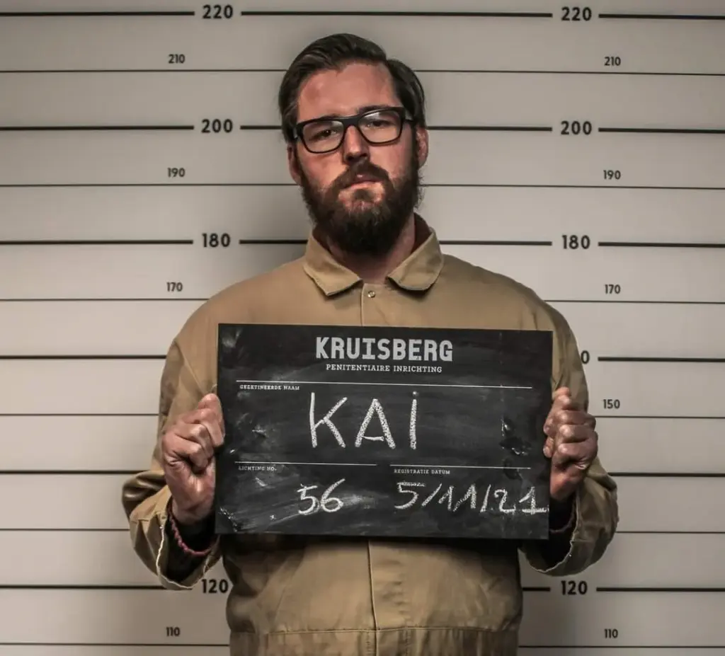 Gijs in de rol van een gevangene van penitentiaire inrichting Kruisberg. Hij houd een bord omhoog met de naam Kai erop geschreven.